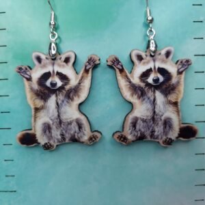 Pair of lightweight 1.5-inch raccoon earrings made from MDF, featuring raised-arm raccoon designs on hypoallergenic hooks, shown against a teal gradient background with measurement markings.